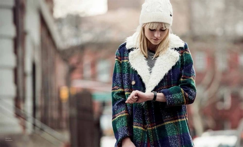 Young woman in a patterned coat and white knit hat checks her watch outdoors against a blurred urban street background.  