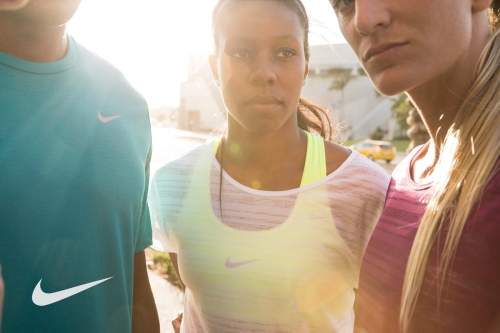 Close-up of three diverse athletes in sportswear with Nike logos, standing outdoors with bright, natural sunlight and urban background.