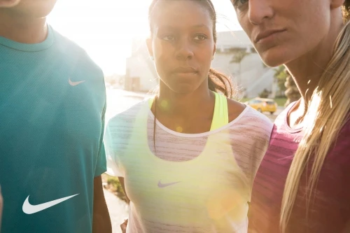 Close-up of three diverse athletes in sportswear with Nike logos, standing outdoors with bright, natural sunlight and urban background.