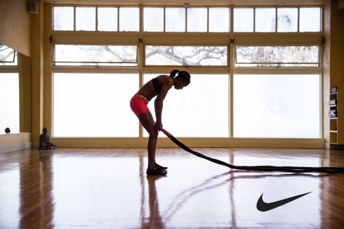 Athletic Black woman in workout gear exercising with battle ropes in a brightly lit gym with a polished wood floor and large windows.