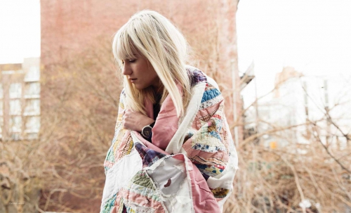 Blonde woman wrapped in a colorful patchwork quilt standing outdoors with buildings and bare trees in the background.