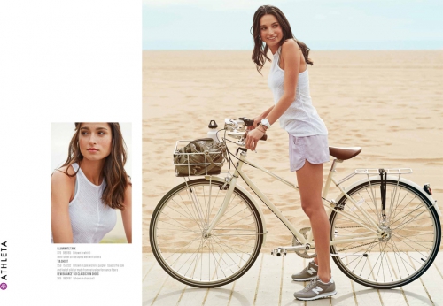Athleta activewear fashion shoot featuring a woman in a white top and lilac shorts posing with a bicycle on a beach boardwalk.