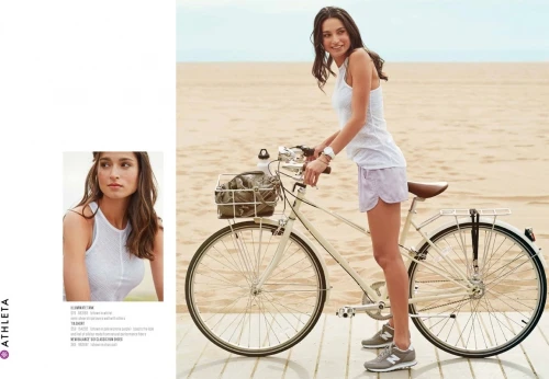 Athleta activewear fashion shoot featuring a woman in a white top and lilac shorts posing with a bicycle on a beach boardwalk.