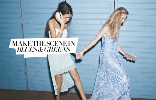 Two women walking together, wearing blue and white dresses with a blue corrugated metal background.  Hand-in-hand, fashion forward.