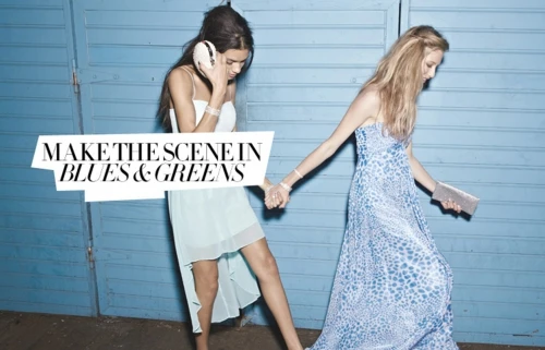 Two women walking together, wearing blue and white dresses with a blue corrugated metal background.  Hand-in-hand, fashion forward.