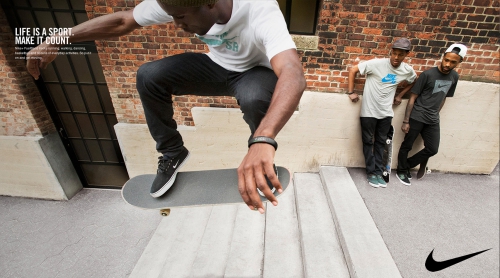 Male skateboarder in mid-air, photographed at a low angle with two men leaning against a brick wall in the background on a sunny day.