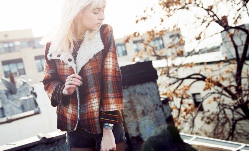 Blonde woman wearing a plaid jacket posing on a rooftop with buildings and trees in the background, creating a fashion editorial shot.