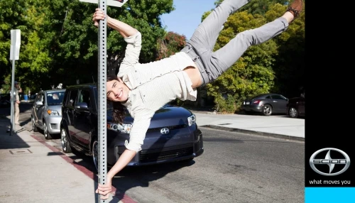 Young man hanging upside down from a street sign on a city street with parked cars and trees in the background in bright sunlight.