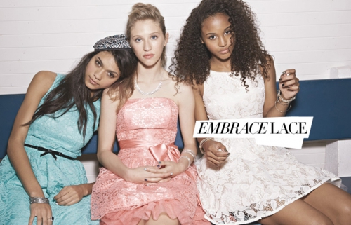 Three young women in lace dresses pose together. One has a bandana, a second wears a statement necklace, and the third has curly hair.