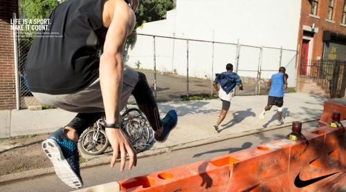 Athlete jumps over barrier on urban street during workout, with two other men running in the background, Nike logo on the barrier.