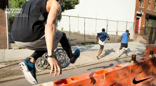 Athlete jumps over barrier on urban street during workout, with two other men running in the background, Nike logo on the barrier.