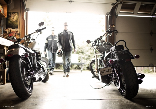 Two motorcycles are parked in a garage, with two adult males walking toward them in the background; sunlight is streaming in.