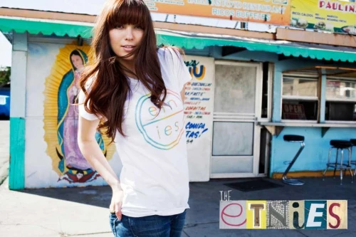 A woman with long brown hair and bangs poses in front of a colorful mural and outdoor eatery wearing a graphic t-shirt and jeans.