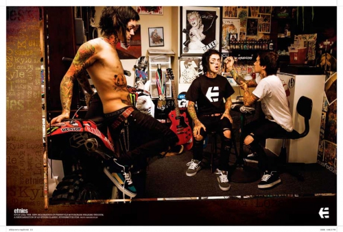 Tattooed man on motorcycle flanked by two other men with tattoos in casual streetwear inside a tattoo parlor-like setting.