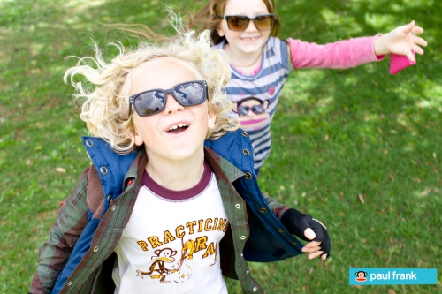 Two smiling children, one boy and one girl, wear sunglasses and play on a grassy lawn in bright sunlight with trees in the background.