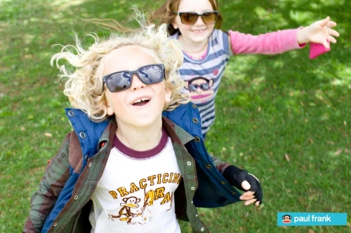 Two smiling children, one boy and one girl, wear sunglasses and play on a grassy lawn in bright sunlight with trees in the background.