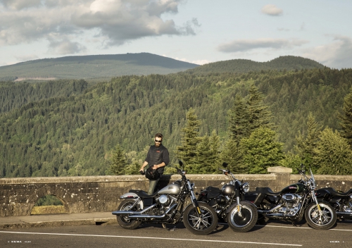 Man standing next to silver motorcycle parked along rural road with mountains and forest in background on a sunny day with some clouds.