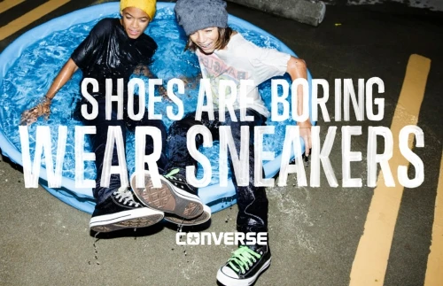 Two young adults pose in a blue plastic pool wearing sneakers, with a Converse logo and the phrase SHOES ARE BORING. WEAR SNEAKERS.