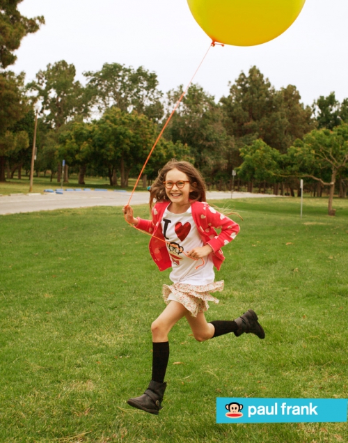 A young girl with glasses joyfully runs across a grassy park with a yellow balloon. Wearing a 'I heart monkeys' t-shirt and skirt.