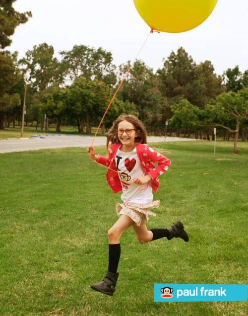 A young girl with glasses joyfully runs across a grassy park with a yellow balloon. Wearing a 'I heart monkeys' t-shirt and skirt.