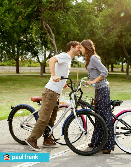 A romantic couple shares a sweet moment outdoors with bicycles in a park setting with lush green trees and warm, natural lighting.