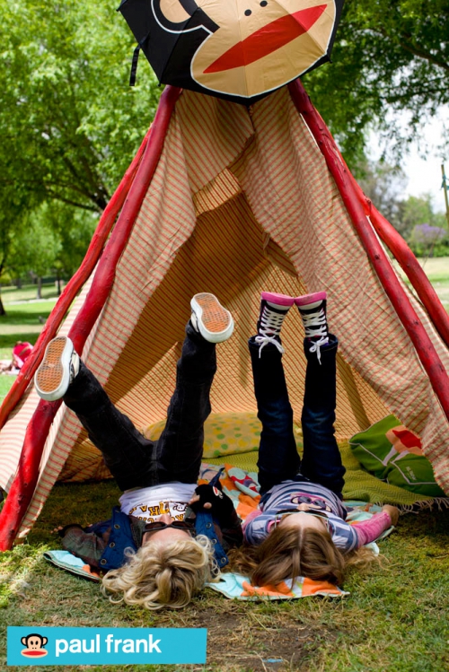 Two children lie upside down on a blanket outside a tent with a cartoon monkey on top, enjoying a playful day in a grassy yard.