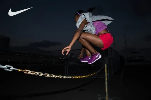 Athletic woman leaps over chain in outdoor nighttime workout, wearing activewear and purple sneakers with the Nike logo in dark setting.