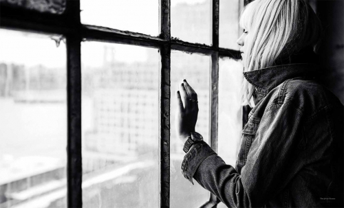 Black and white portrait of a woman looking out a multi-pane grid window wearing a denim jacket.