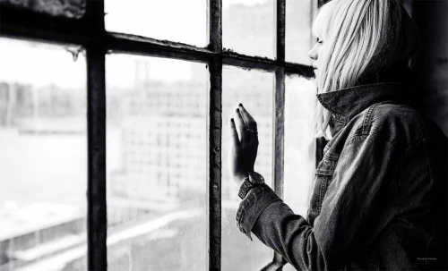 Black and white portrait of a woman looking out a multi-pane grid window wearing a denim jacket.