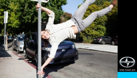 Man spinning horizontally around a metal signpost on a sunny street with parked cars and a vertical branding panel on the right.