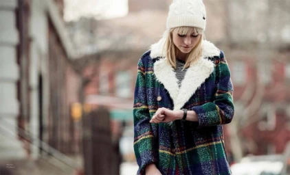 Woman wearing plaid coat and white beanie looking at her watch on city street