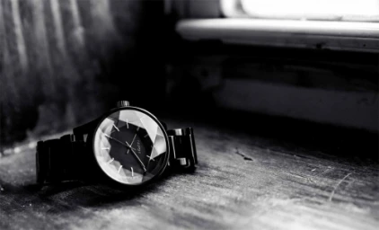 Close-up monochrome photograph of a faceted black wristwatch resting on a textured wooden surface near a window.