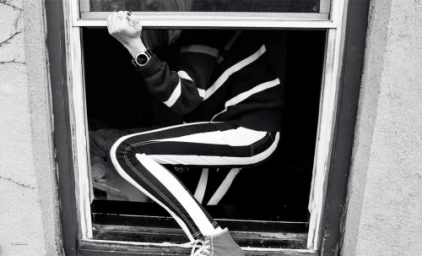 Black and white portrait of a person crouching inside a window frame wearing striped pants and holding the frame ledge