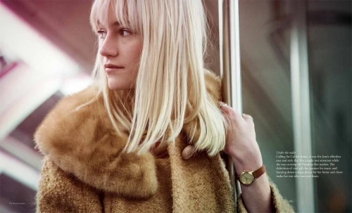 Blonde woman with bangs wearing a brown faux fur collared coat holding a metal pole inside public transit.