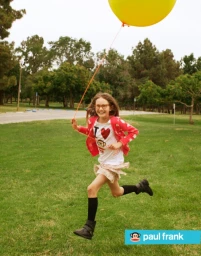 Young child running across a grassy park holding the string of a large yellow balloon, photographed by Rennie Solis.