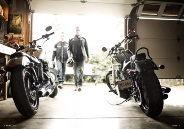 Two motorcycles parked in the foreground of an open garage with two men walking in from daylight.