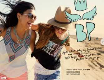 Two smiling young women walking together on a sandy beach wearing casual festival fashion with sunglasses and a straw hat.