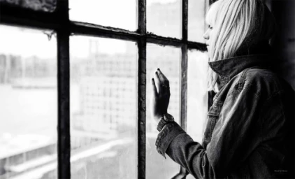 Black and white portrait of a woman looking out a multi-pane grid window wearing a denim jacket.