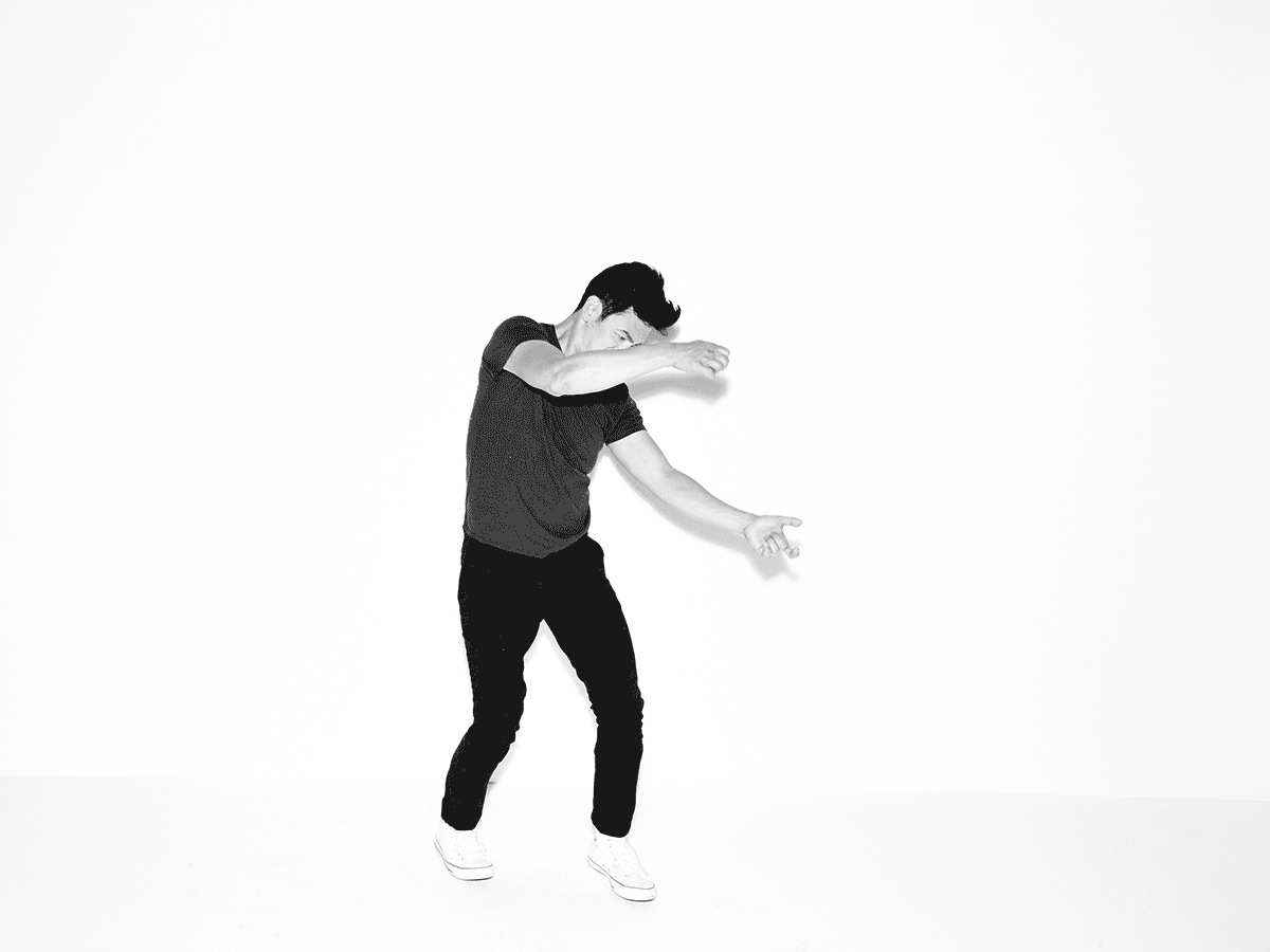 Black and white image of a male dancer in motion, wearing dark clothing and light sneakers, set against a stark white background.