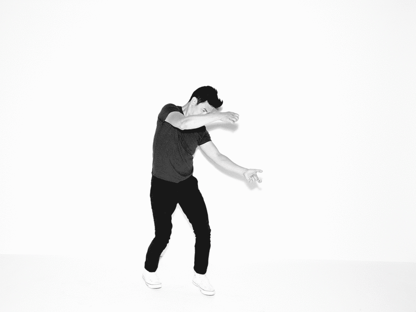 Black and white image of a male dancer in motion, wearing dark clothing and light sneakers, set against a stark white background.