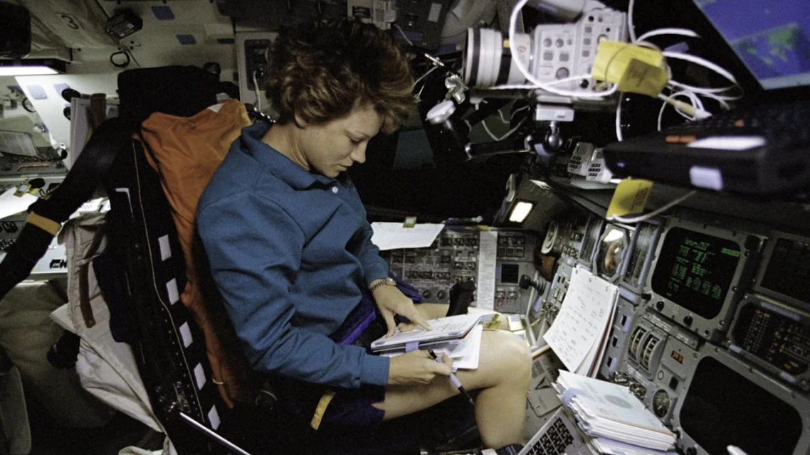 Astronaut working on the space shuttle, seated amidst instrument panels and equipment, writing in a notebook, dressed in blue shirt and shorts.