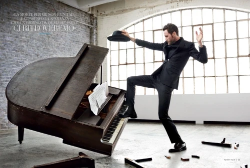Adult male in a dark suit stands on a piano with sheet music in a bright indoor space with exposed brick and large windows.