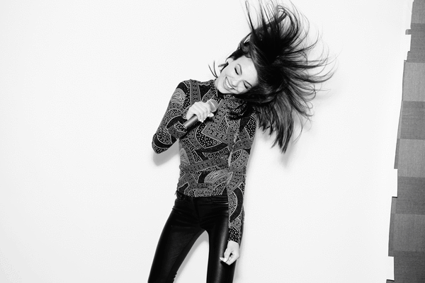 Energetic studio portrait of a young adult female singer with long dark hair, holding a microphone and smiling against a white backdrop.
