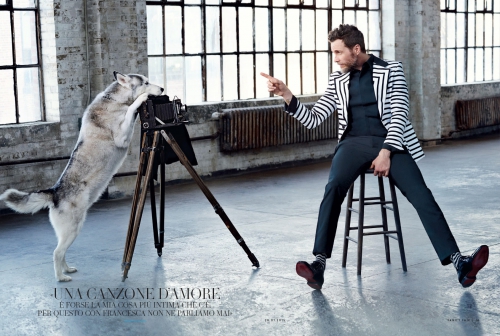 Editorial fashion shot of an adult male in striped blazer pointing his finger and a dog leaning against an antique camera indoors.