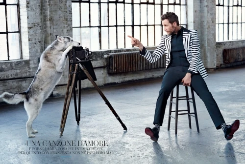 Editorial fashion shot of an adult male in striped blazer pointing his finger and a dog leaning against an antique camera indoors.