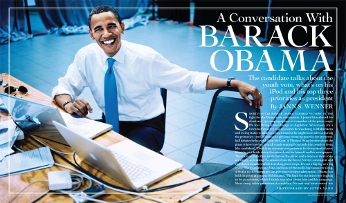 Barack Obama, wearing a white shirt and blue tie, is photographed smiling at a desk with open laptops in a naturally lit indoor setting.
