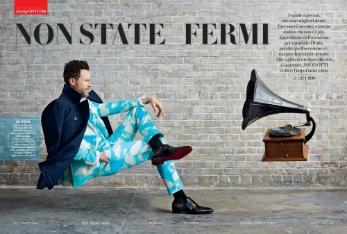 Man sits in a cloud-patterned suit, posing with vintage phonograph against a gray brick wall, embodying retro style and modern fashion.
