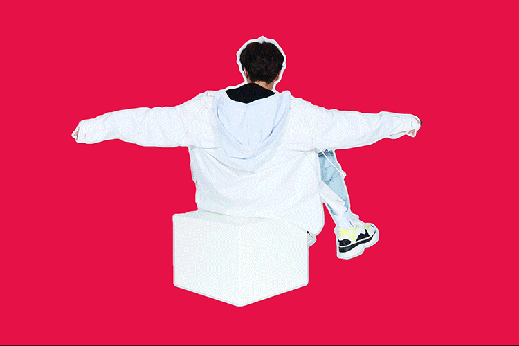 Back view of person in white jacket and blue jeans sitting on a white block against a red background with arms outstretched.