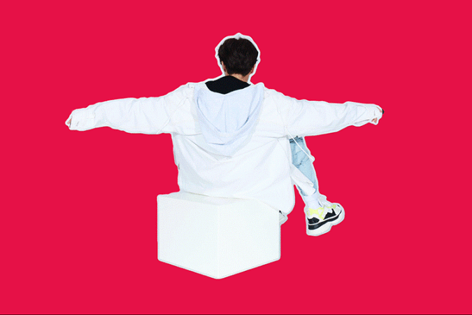 Back view of person in white jacket and blue jeans sitting on a white block against a red background with arms outstretched.