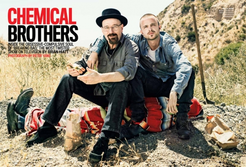 Two actors from 'Breaking Bad' sit on a blanket outdoors. One wears a hat; the setting includes a shovel, gravel, and hills.